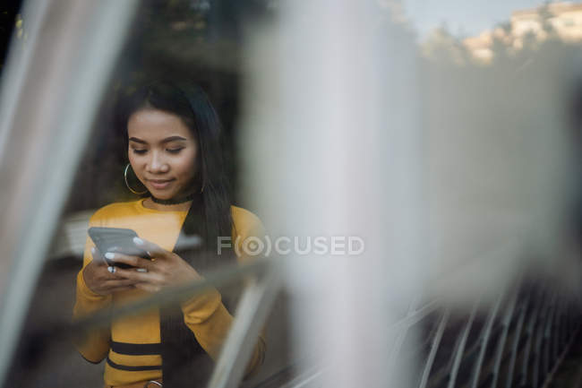 Elegante graziosa sorridente donna asiatica navigando telefono cellulare su strada vicino alla costruzione di metallo — Foto stock