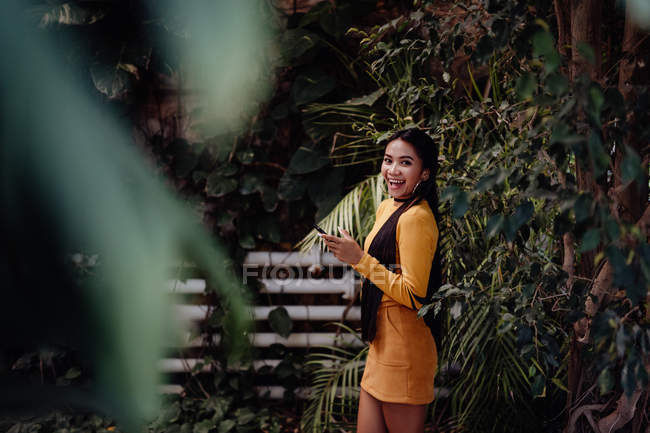 Побічний вид стильного посміхаючись брюнетку азіатська жінка серфінг смартфон на фоні стіни з альпіністськими зеленими рослинами — стокове фото