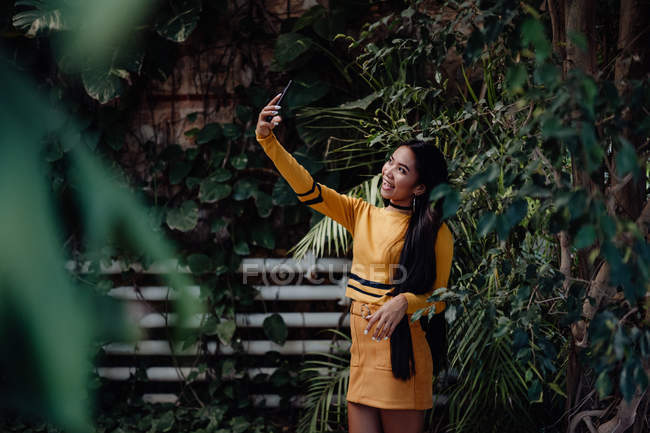 Вид сбоку на стильную улыбчивую брюнетку азиатской женщины, серфингующей смартфон на фоне стены с лазающими зелеными растениями — стоковое фото
