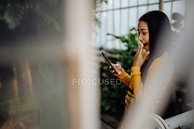 Vista laterale di elegante bruna sorridente smartphone donna asiatica surf sullo sfondo della parete con arrampicata piante verdi — Foto stock