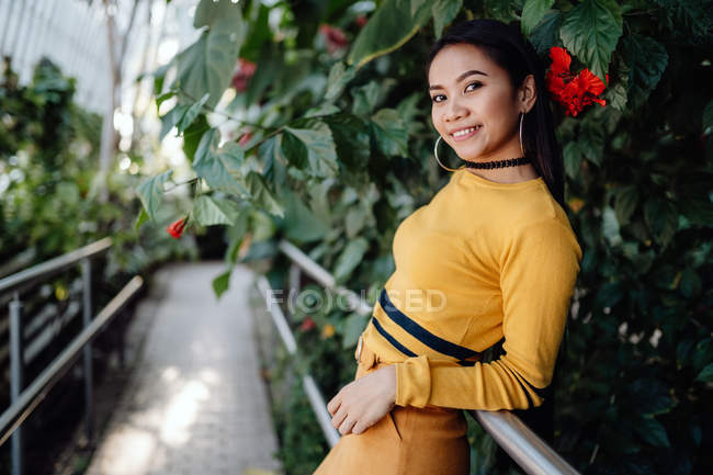 Side view of Asian woman leaning on metal fence with climbing plants and looking at camera — Stock Photo