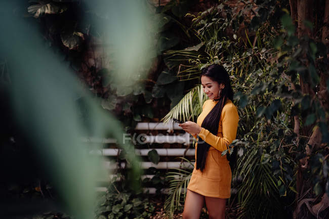 Вид сбоку на стильную улыбчивую брюнетку азиатской женщины, серфингующей смартфон на фоне стены с лазающими зелеными растениями — стоковое фото