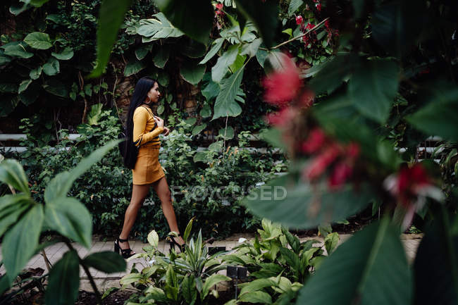 Vista lateral de la mujer asiática de moda con pelo largo y oscuro en camisa amarilla y falda corta caminando en hermoso jardín y mirando hacia otro lado - foto de stock