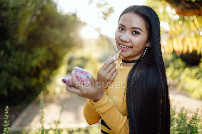 Charmante trendige Asiatin mit langen dunklen Haaren im gelben Hemd blickt in die Kamera und setzt Lipgloss in den sonnigen Park — Stockfoto