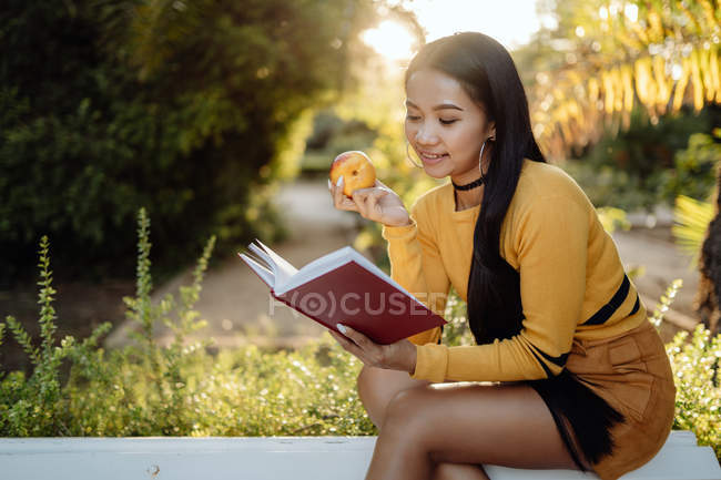 Brunette Asian woman reading book in red cover and eating tasty apple sitting on white bench in park — Stock Photo