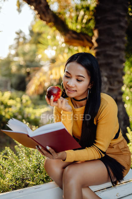 Brunette Asian woman reading book in red cover and eating tasty apple sitting on white bench in park — Stock Photo