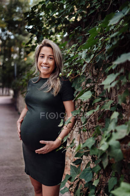 Glücklich schwangere Frau in legerem Kleid streichelt Bauch, während sie auf Gehweg in Park mit grünen Bäumen steht — Stockfoto