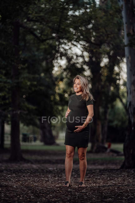 Mujer embarazada feliz en vestido casual acariciando el vientre mientras está de pie en el camino en el parque con árboles verdes - foto de stock
