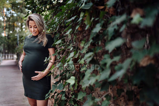 Счастливая беременная женщина в повседневной одежде гладит живот, стоя на дорожке в парке с зелеными деревьями — стоковое фото