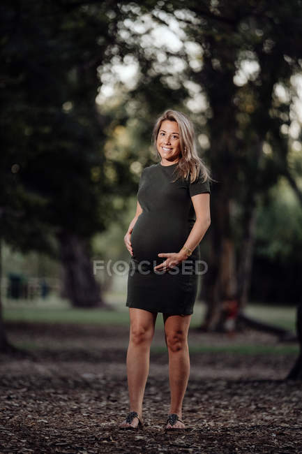 Heureuse femme enceinte en robe décontractée caressant le ventre tout en se tenant sur le chemin dans le parc avec des arbres verts dans la journée ensoleillée d'été — Photo de stock