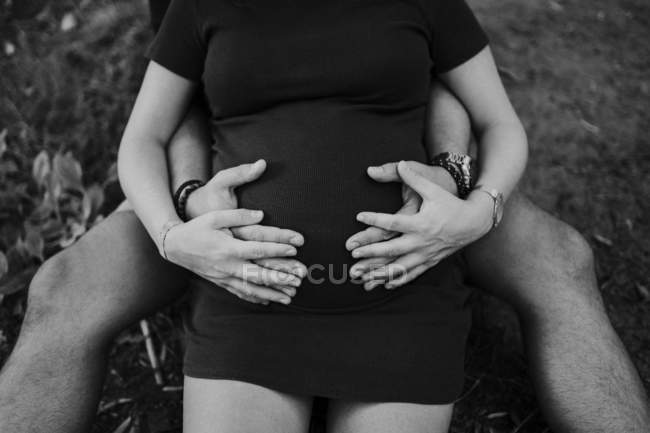 Обрізаний чоловік обіймає вагітну дружину і обидва тримають руки на животі, сидячи разом на траві в парку — стокове фото