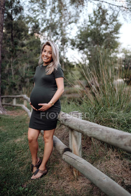 Schöne schwangere Frau in lässigem Kleid, lächelnd und Händchen haltend auf dem Bauch, während sie neben Holzbrücke steht — Stockfoto
