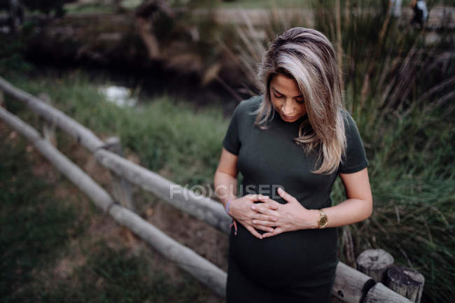 Belle femme enceinte en robe décontractée souriante et tenant la main sur le ventre tout en se tenant à côté du pont en bois — Photo de stock