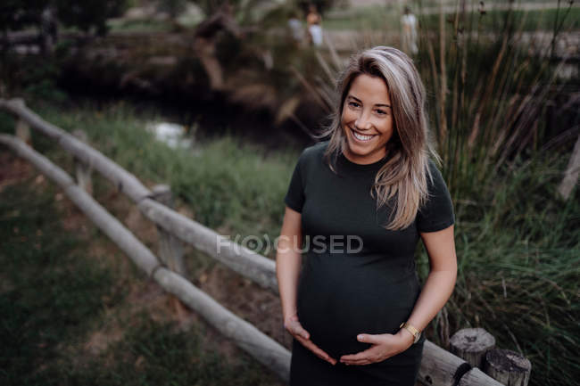 Красивая беременная женщина в повседневном платье улыбается и держит руки на животе, стоя рядом с деревянным мостом — стоковое фото