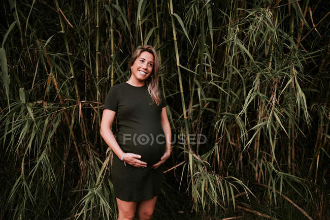 Счастливая беременная женщина в повседневной одежде гладит живот, стоя на зеленой стене деревьев в солнечный летний день с голубым небом — стоковое фото
