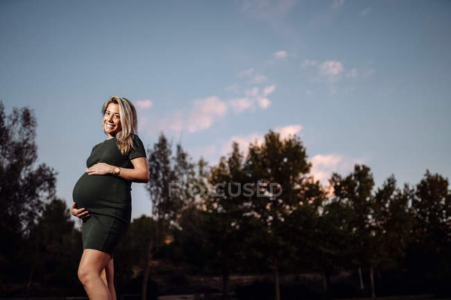 Heureuse femme enceinte en robe décontractée caressant le ventre tout en se tenant sur le chemin dans le parc avec des arbres verts — Photo de stock