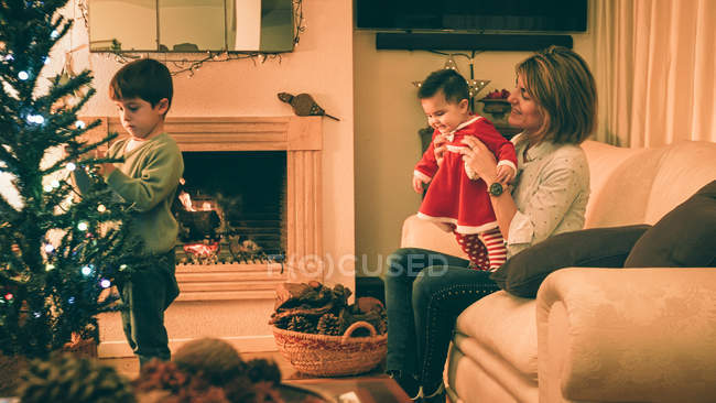 Madre jugando con el bebé mientras el hijo decora el árbol de Navidad - foto de stock