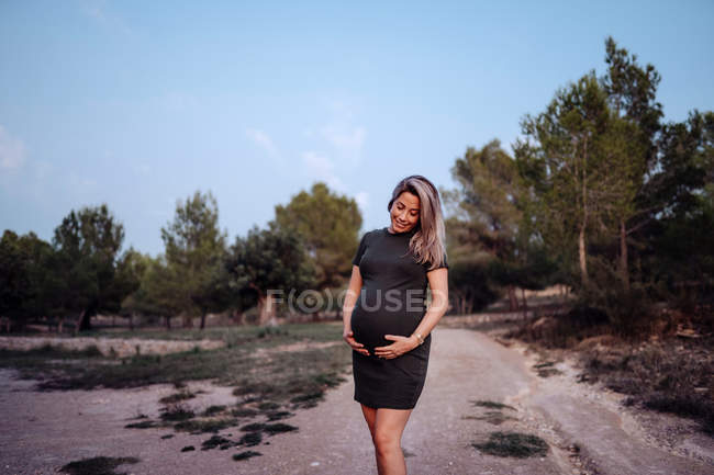 Happy pregnant woman in casual dress stroking belly while standing on pathway in park with green trees in sunny summer day — Stock Photo