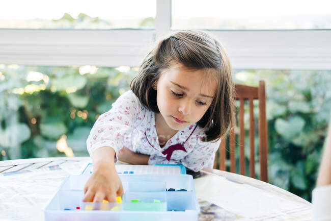 Adorable niña concentrada poniendo pequeños bloques de colores para abordar y crear mosaico mientras juega juego educativo - foto de stock