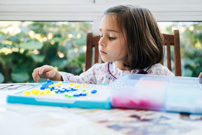 Adorable niña concentrada poniendo pequeños bloques de colores para abordar y crear mosaico mientras juega juego educativo - foto de stock