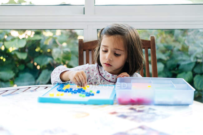 Приваблива сконцентрована дівчинка кладе маленькі кольорові блоки на борт і створює мозаїку, граючи в навчальні ігри. — Stock Photo