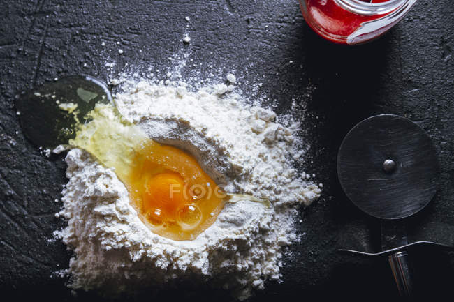 Верхний вид разбитого яйца в муке на текстурированной черной поверхности с круглым ножом и стеклянным горшком — стоковое фото