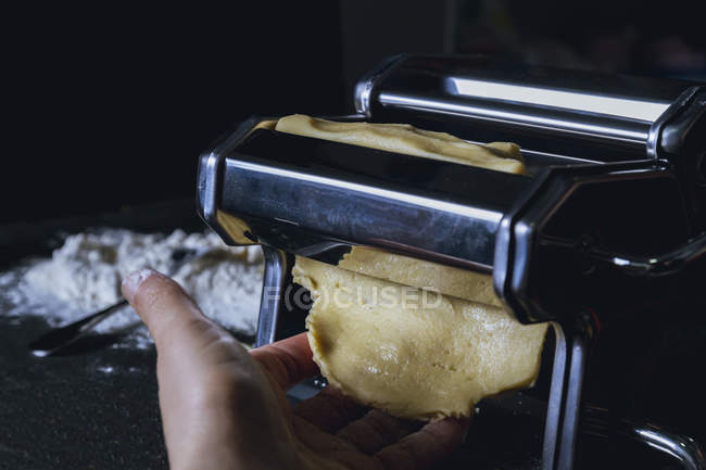 Primer plano de la mano de la persona rodando masa a través de la máquina de pasta mientras se prepara pasta casera fresca en la mesa - foto de stock