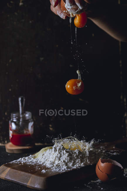 Mano de la persona rompiendo huevos sobre harina mientras prepara la masa en la mesa con cáscara de huevo y maceta de vidrio - foto de stock