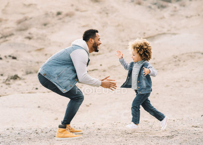 Cheerful ethnic stylish man playing with curly happy toddler on sandy hills at daytime — Stock Photo