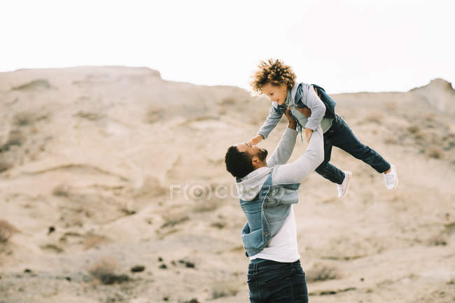 Cheerful ethnic stylish man lifting and playing with curly happy toddler on sandy hills at daytime — Stock Photo