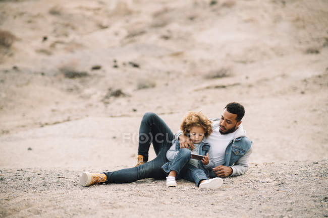 African American casual man leaning on elbow and looking at curly toddler sitting beside with mobile phone — Stock Photo