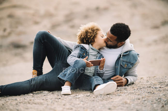 African American casual man leaning on elbow and kissing curly toddler daughter sitting beside with mobile phone — Stock Photo