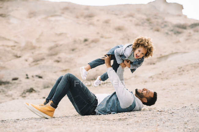 Cheerful ethnic man playing with curly happy ethnic toddler on bright sandy hills at daytime — Stock Photo