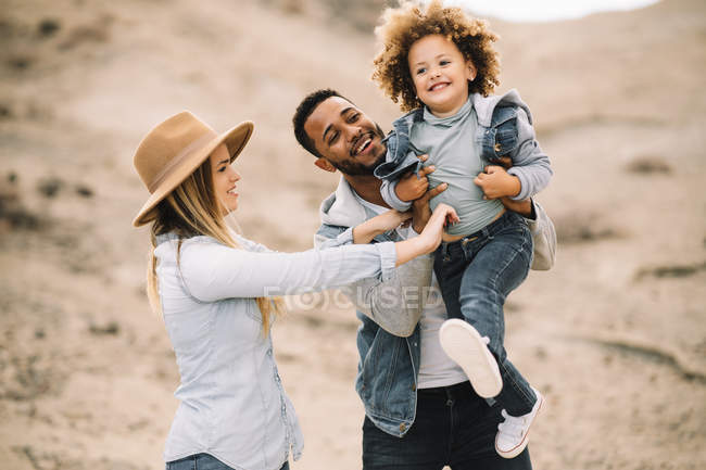 Веселые многорасовые родители держат в руках очаровательного кудрявого этнического малыша и веселятся на песчаном ландшафте — стоковое фото