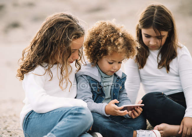 Konzentrierte, ungezwungene Mixed Race Kids, die es sich auf Sand gemütlich machen und tagsüber ihr Handy teilen — Stockfoto