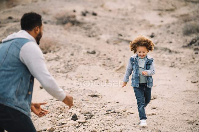 Cheerful ethnic man playing with curly happy ethnic toddler on bright sandy hills at daytime — Stock Photo