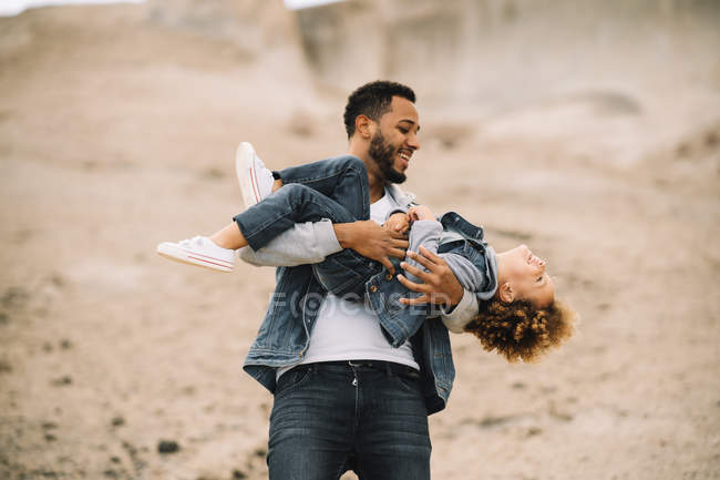 Homem barbudo alegre em roupas elegantes segurando jogar como carregando criança étnica feliz no deserto durante o dia — Fotografia de Stock
