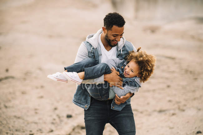 Homem barbudo alegre em roupas elegantes segurando jogar como carregando criança étnica feliz no deserto durante o dia — Fotografia de Stock