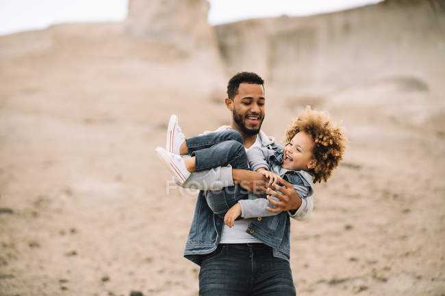 Cheerful bearded man in stylish clothes holding playing as carrying happy ethnic toddler in desert at daytime — Stock Photo