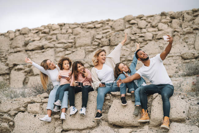 Black man taking selfie with mobile phone with children and women while sitting on stone wall at daytime — Stock Photo