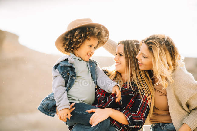 Cheerful mother in checkered shirt holding casual toddler as joyful female friend putting hat on child on nature at daytime — Stock Photo
