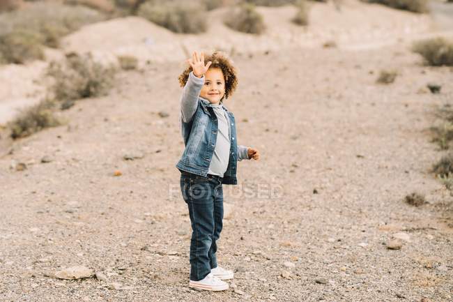 Adorable enfant ethnique bouclé vêtu de vêtements décontractés agitant la main sur fond flou — Photo de stock