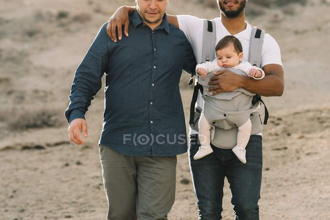 Cropped of casual men holding little calm baby in carrier while strolling on nature at daytime — Stock Photo