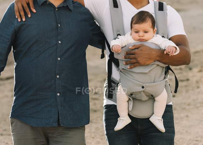 Cropped of casual men holding little calm baby in carrier while strolling on nature at daytime — Stock Photo