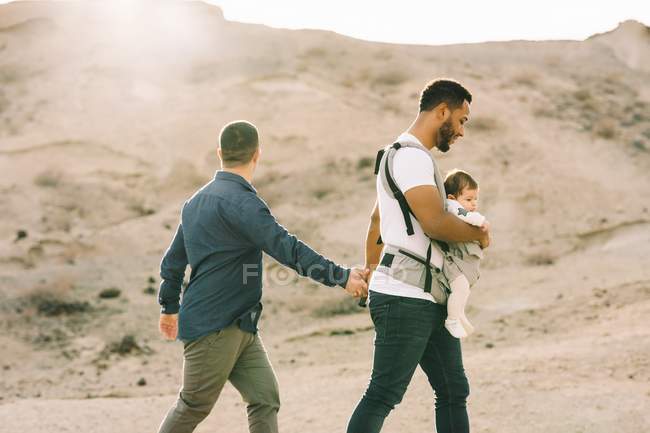 Side view of casual homosexual couple with little calm baby in carrier holding hands while strolling on nature at daytime — Stock Photo