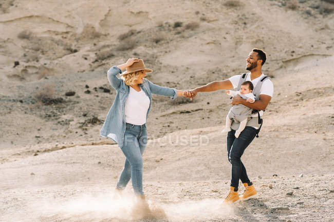 Веселый мужчина, несущий маленького ребенка и держащийся за руки с блондинкой женой, танцуя в песчаной пустыне — стоковое фото