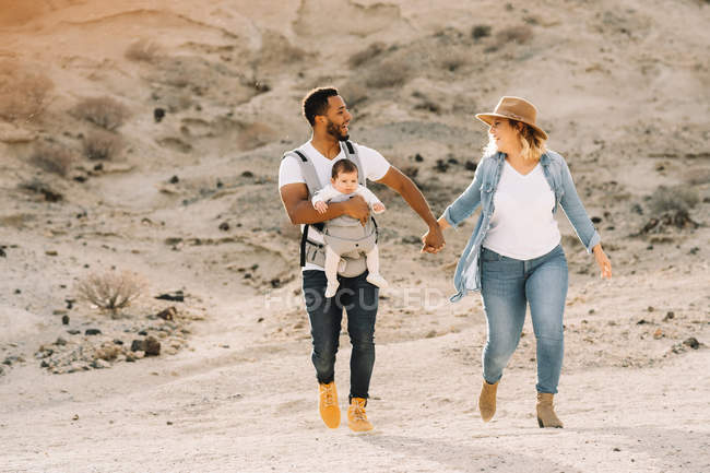 Веселый мужчина, несущий маленького ребенка и держащийся за руки с блондинкой женой во время прогулки по песчаной пустыне — стоковое фото