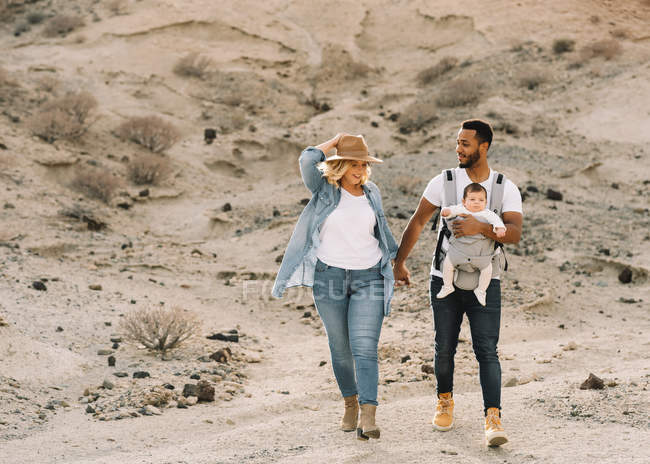 Homem alegre carregando pequeno bebê e de mãos dadas com a esposa loira enquanto caminha no deserto arenoso — Fotografia de Stock