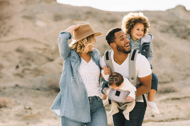 Feliz esposa caucásica casual y negro sonriente marido llevando niños en la naturaleza durante el día - foto de stock