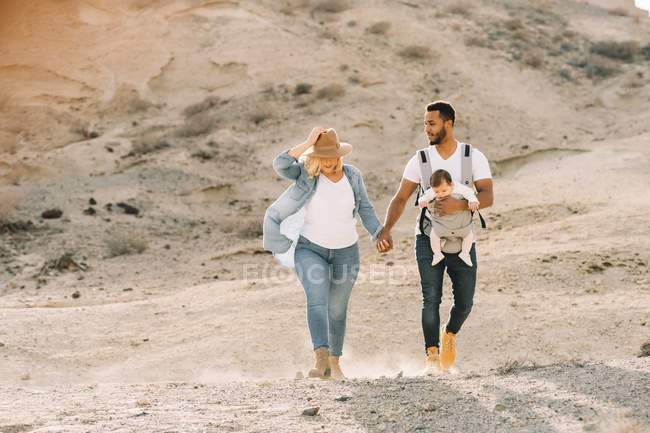 Cheerful man carrying little baby and holding hands with blonde wife while walking in sandy desert — Stock Photo
